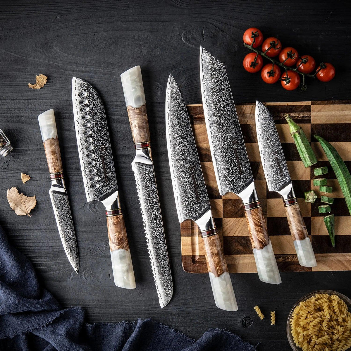 Damascus kitchen knives with wooden and resin handles on dark wood, beside cherry tomatoes and pasta