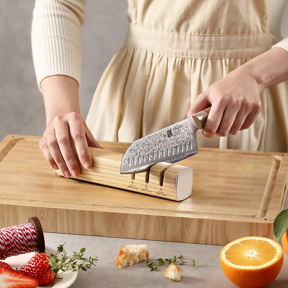 Person sharpening a Damascus kitchen knife on a wooden sharpener with orange and strawberries nearby